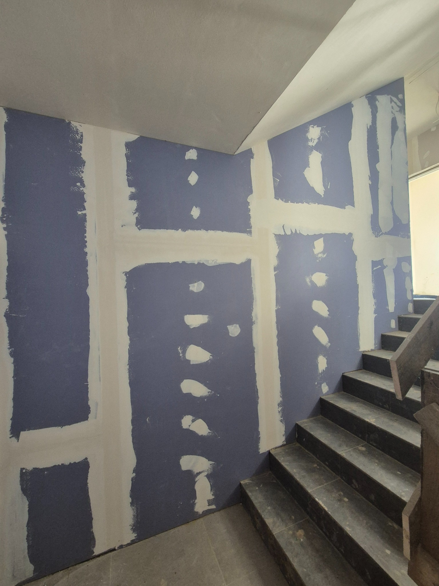 Ściana z niebieską płytą gipsowo-kartonową, częściowo pokryta szpachlą, widoczne łączenia i ślady po wkrętach, obok schody z ciemnego kamienia.