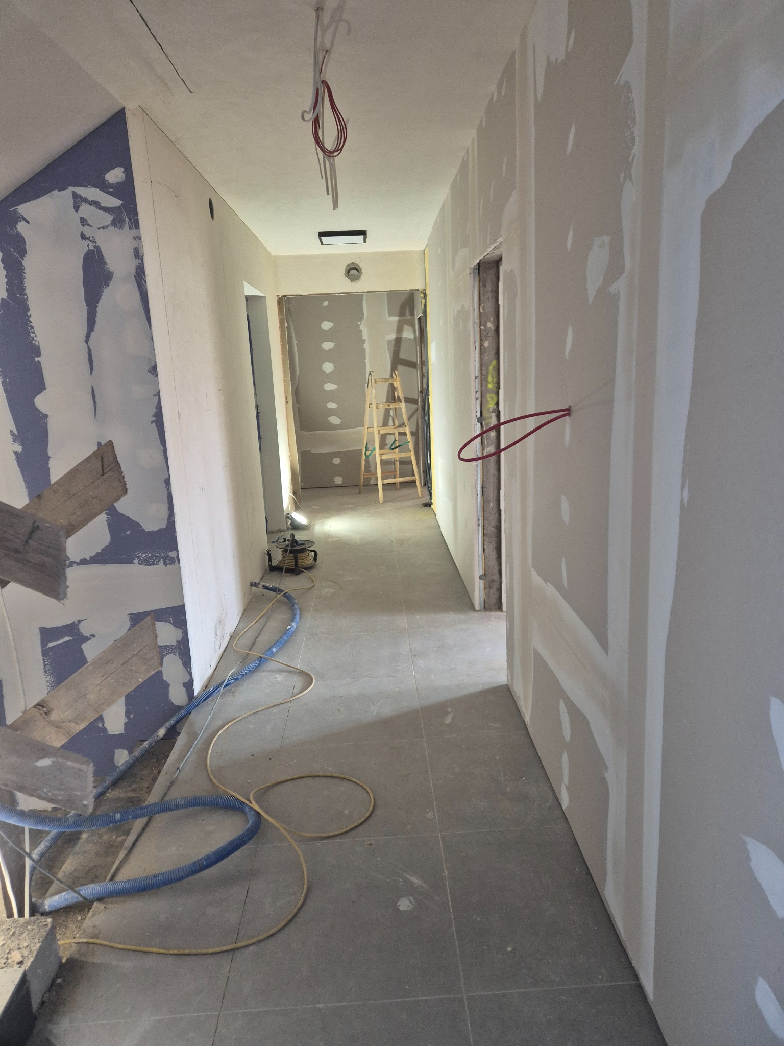 Korytarz w trakcie remontu z widocznymi ścianami z płyt kartonowo-gipsowych, szpachlowanymi łączeniami, kable wystające ze ściany, drabina i narzędzia na podłodze.