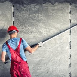 Usługi Budowlane Łukasz Chyłkowski - Tynkarz w czerwonym kombinezonie wygładza szarą, surową ścianę długą łatą. Widoczne listwy prowadzące. Praca w toku.