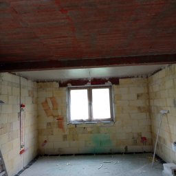 Usługi Budowlane Łukasz Chyłkowski - Wnętrze w trakcie prac przygotowawczych: surowy strop z czerwonymi śladami, ściany z pustaków z widocznymi instalacjami, okno z naturalnym światłem.