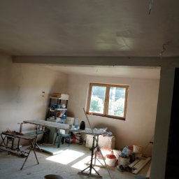 Usługi Budowlane Łukasz Chyłkowski - Wnętrze pokoju po nałożeniu gładzi, widoczne ślady po taśmach malarskich na suficie, okno z widokiem na zieleń, wiadra i narzędzia malarskie.