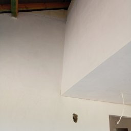 Usługi Budowlane Łukasz Chyłkowski - Wnętrze po pracach wykończeniowych: gładkie, jasne ściany i sufit w nowym budynku. Widoczne drewniane elementy konstrukcyjne pod dachem i otwór w ścianie.