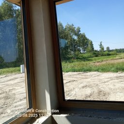 Usługi Budowlane Łukasz Chyłkowski - Wnętrze w trakcie prac wykończeniowych, widok przez okno na zewnątrz. Ściany przygotowane do dalszych prac, widoczne ślady po obróbce.