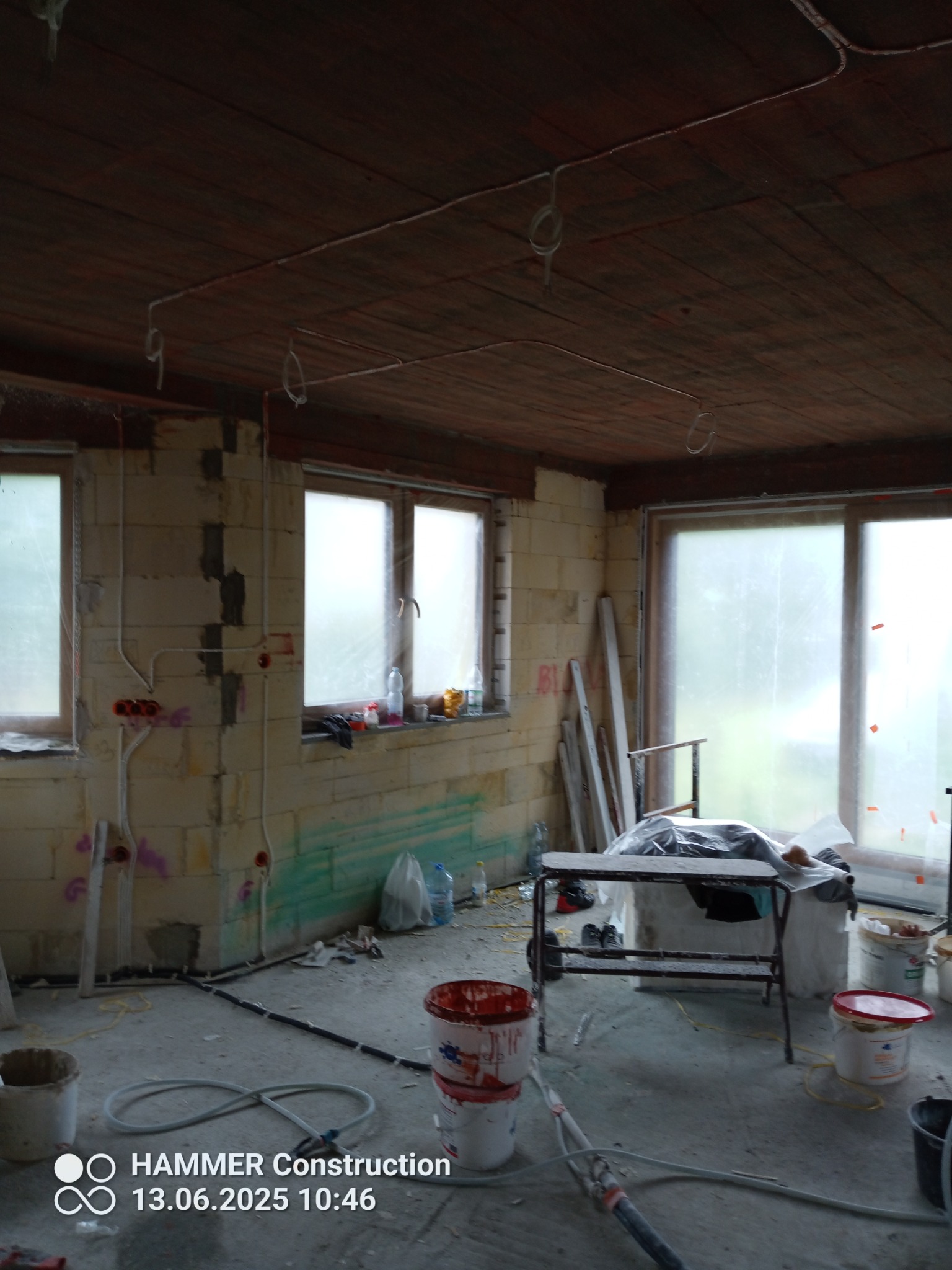 Pokój w trakcie remontu: surowe ściany z instalacjami elektrycznymi, drewniany sufit z przewodami, okna zasłonięte folią, widoczne narzędzia i materiały budowlane.