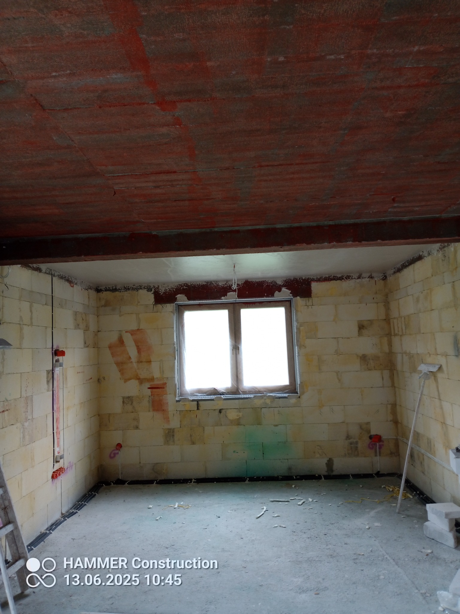 Wnętrze w trakcie prac przygotowawczych: surowy strop z czerwonymi śladami, ściany z pustaków z widocznymi instalacjami, okno z naturalnym światłem.