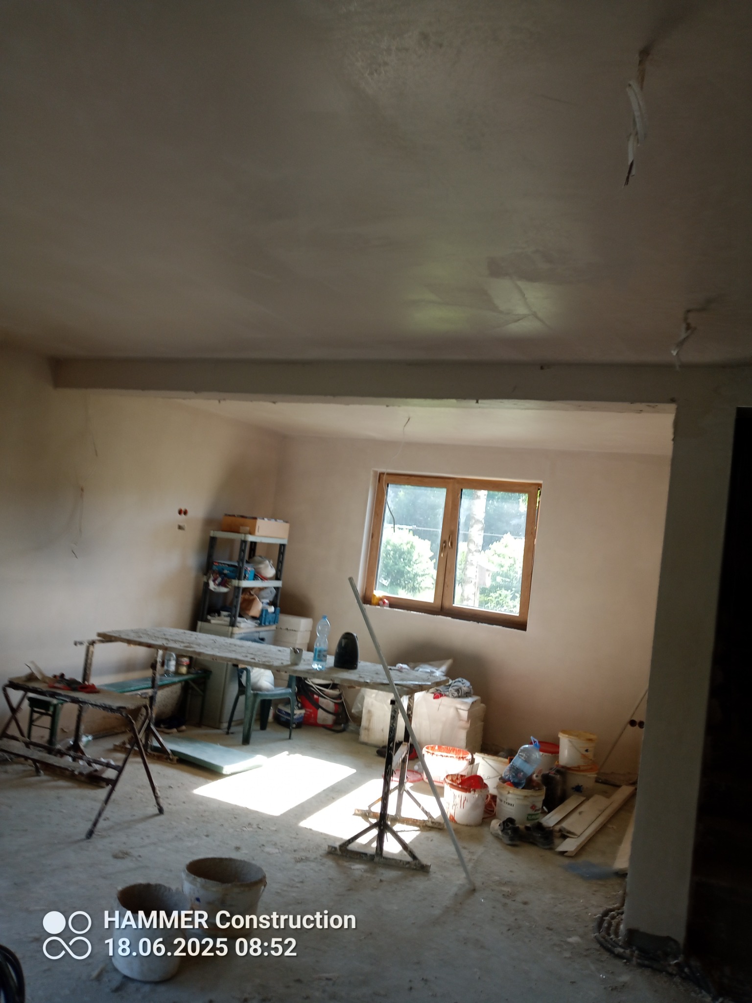 Wnętrze pokoju po nałożeniu gładzi, widoczne ślady po taśmach malarskich na suficie, okno z widokiem na zieleń, wiadra i narzędzia malarskie.