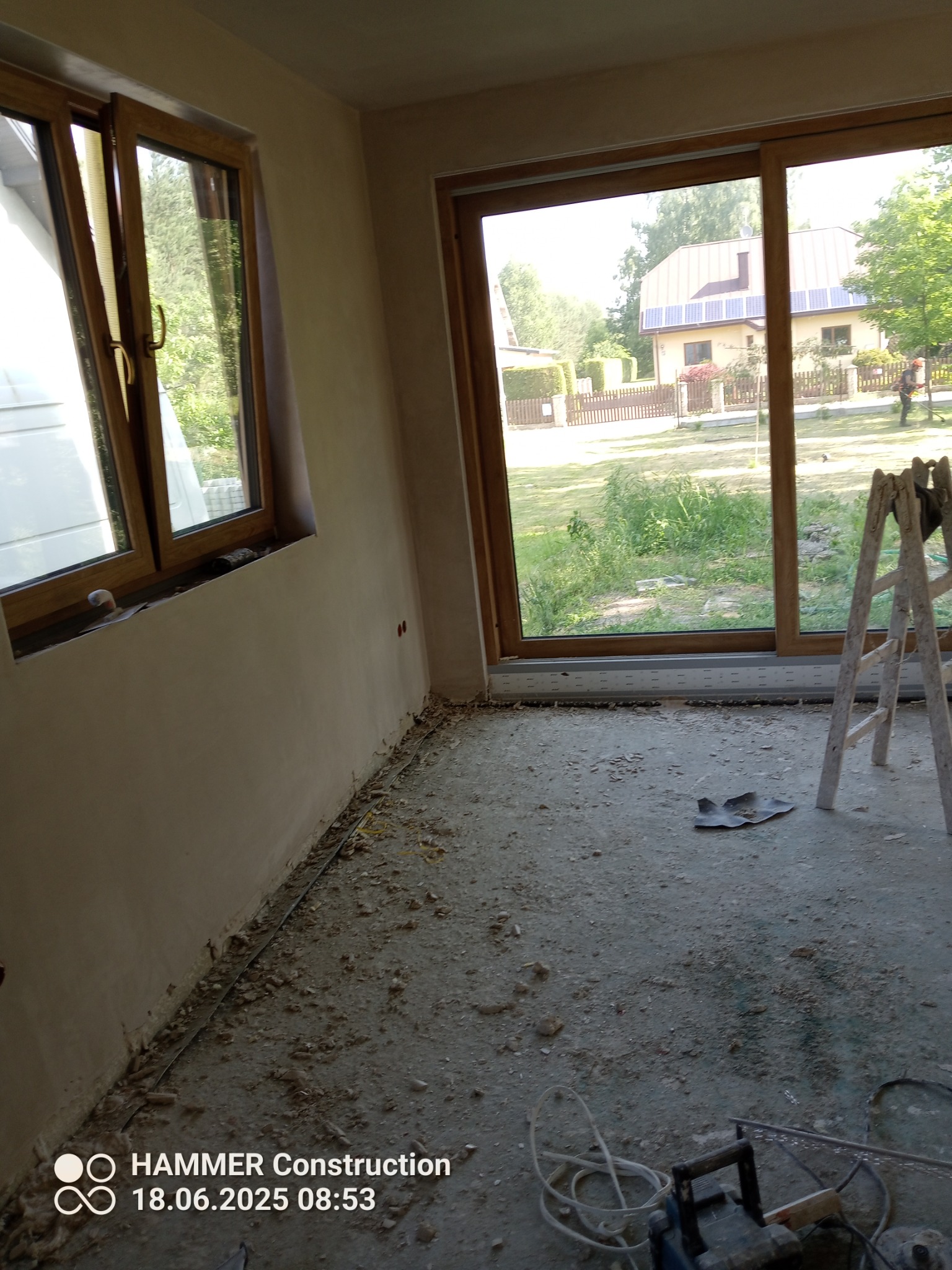 Wnętrze w trakcie prac tynkarskich: ściany pokryte świeżym tynkiem, widoczne okna z drewnianą ramą, drabina i narzędzia na podłodze. Dom z panelami słonecznymi w tle.