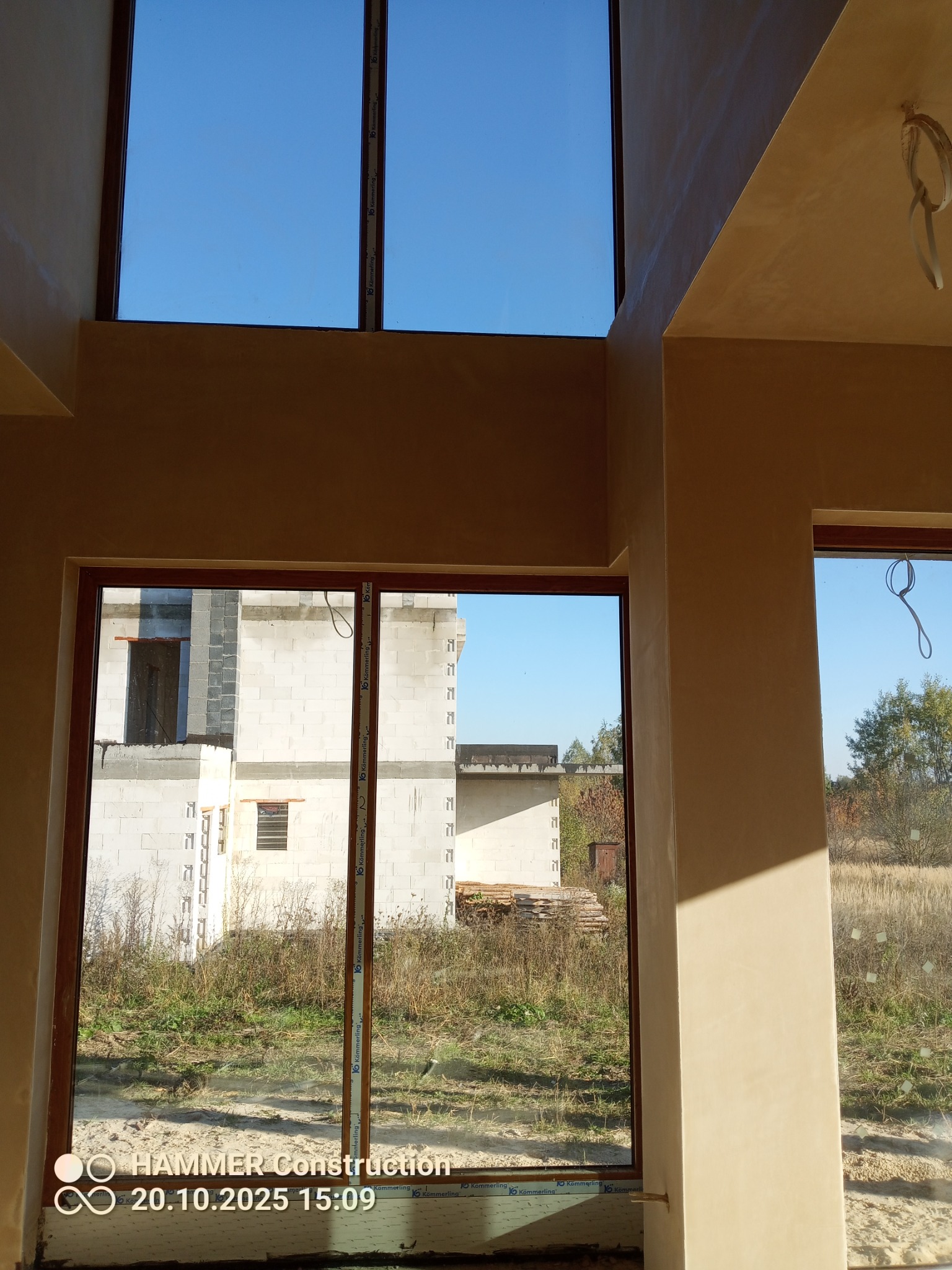 Jasne wnętrze w trakcie wykańczania: ściany pokryte beżową warstwą, duże okna z widokiem na budynek w budowie i błękitne niebo. Widoczne detale wykończeniowe.