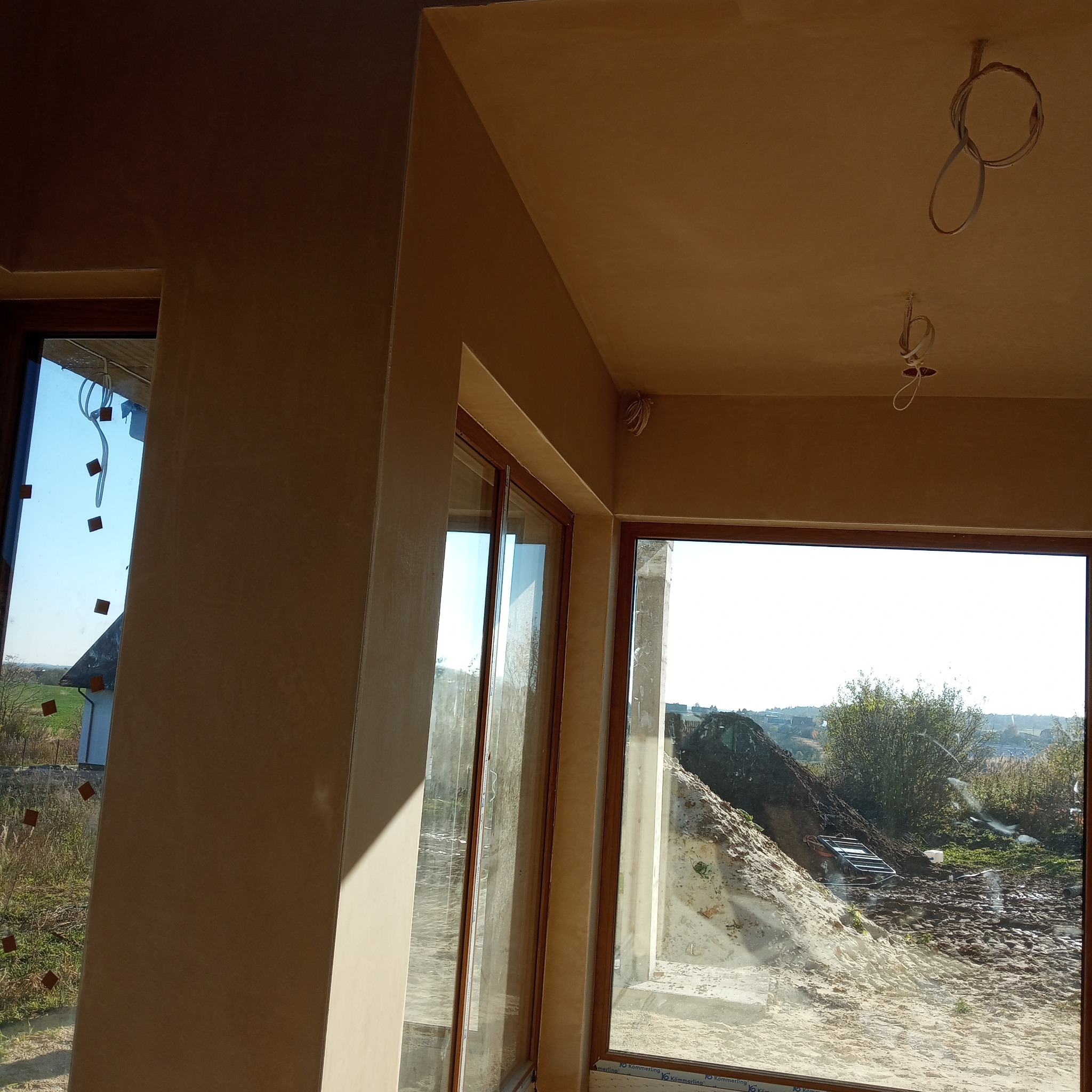 Jasne wnętrze w trakcie prac wykończeniowych. Ściany i sufit pokryte świeżo nałożoną, beżową warstwą. Widoczne okna z widokiem na zewnątrz i wystające kable.