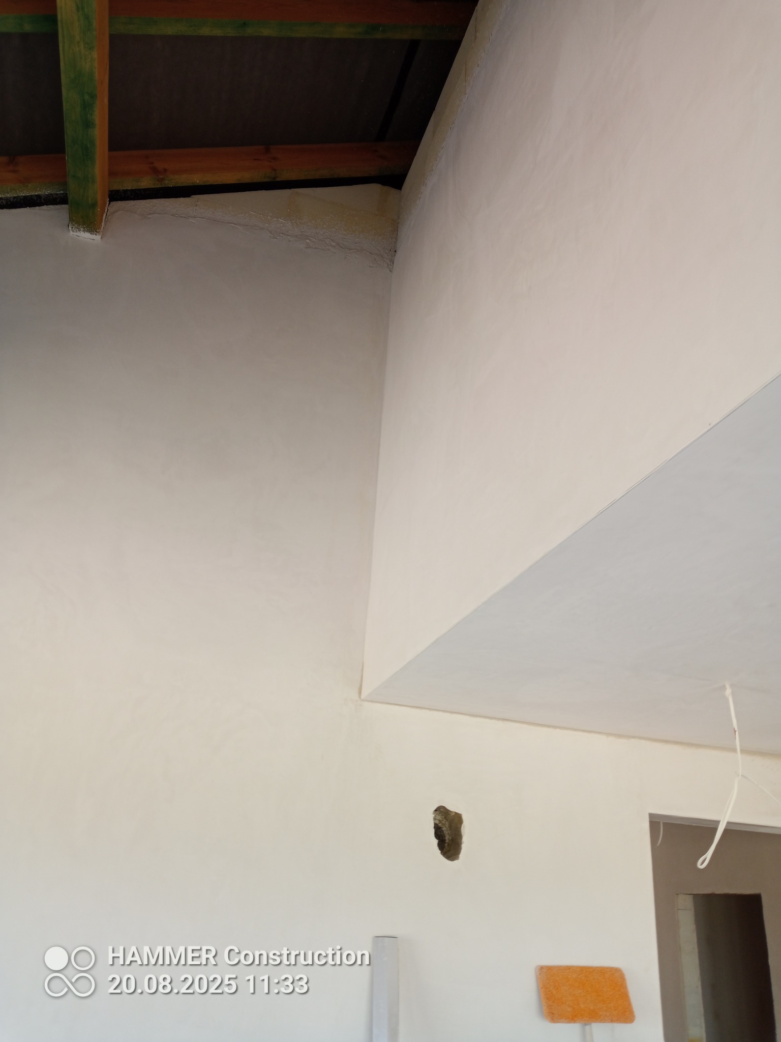 Wnętrze po pracach wykończeniowych: gładkie, jasne ściany i sufit w nowym budynku. Widoczne drewniane elementy konstrukcyjne pod dachem i otwór w ścianie.