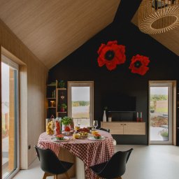 Mariusz Buja - Nowoczesne wnętrze domu z bali: drewniane ściany i sufit, czarna ściana z dekoracjami kwiatowymi, stół z czerwonym obrusem i czarne krzesła.