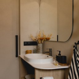 Mariusz Buja - Nowoczesna łazienka z umywalką nablatową, lustrem o zaokrąglonym kształcie i beżową kolorystyką. Elementy dekoracyjne: suszone kwiaty i minimalistyczne akcesoria.