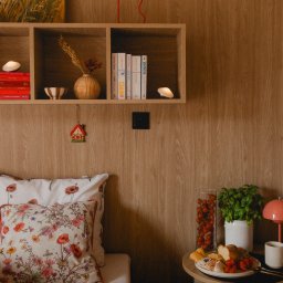 Mariusz Buja - Przytulne wnętrze sypialni z drewnianą ścianą, półką z książkami, lampką i obrazem z motywem maków. Na stoliku nocnym przekąski i roślina w doniczce.