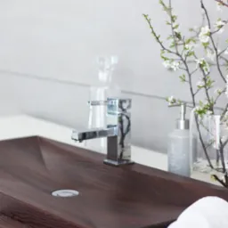 Ciemnobrązowa umywalka z drewna egzotycznego, chromowana bateria, biały ręcznik zwinięty w rulon i gałązka kwitnącej wiśni w tle, minimalistyczny wystr&oacute;j łazienki.