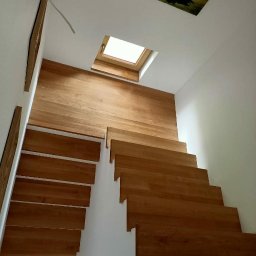 Mariusz Buja - Drewniane schody z jasnego drewna, widok z dołu, z oknem dachowym w tle. Minimalistyczny design, białe ściany, ciepłe oświetlenie. Detal wykończenia wnętrza.