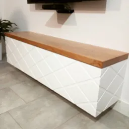 Biała, wisząca szafka RTV z drewnianym blatem i dekoracyjnym frezowaniem w romby, zamontowana na ścianie w nowoczesnym salonie, widok z perspektywy podłogi.