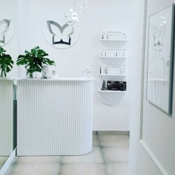 Mariusz Buja - Białe, minimalistyczne wnętrze salonu: lada recepcyjna z roślinami, półki z kosmetykami, dekoracja motyla i grafika na ścianie. Stonowana kolorystyka, nowoczesny design.