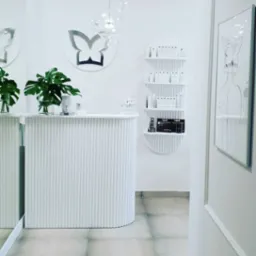 Białe, minimalistyczne wnętrze salonu: lada recepcyjna z roślinami, półki z kosmetykami, dekoracja motyla i grafika na ścianie. Stonowana kolorystyka, nowoczesny design.