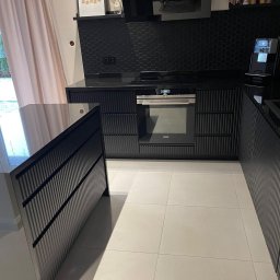 Mariusz Buja - Nowoczesna, czarna kuchnia z frezowanymi frontami, blatem z połyskiem i heksagonalnymi płytkami na ścianie. Widoczny piekarnik Siemens i ekspres do kawy.