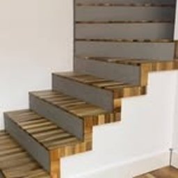 Mariusz Buja - Nowoczesne schody z drewnianymi stopniami o mozaikowej fakturze i szarymi podstopnicami, widok z dołu, na tle białej ściany i listwy przypodłogowej.