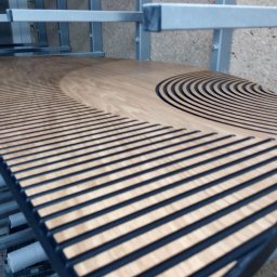 Mariusz Buja - Panel akustyczny z drewna z wyfrezowanymi, równoległymi rowkami i motywem półkola, leżący na metalowym regale w warsztacie. Widok z góry.