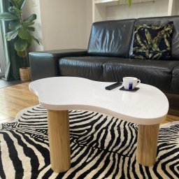 Mariusz Buja - Biały stolik kawowy o nieregularnym kształcie na drewnianych nogach, z filiżanką i pilotem, na tle sofy i dywanu w zebrę. Nowoczesny design.