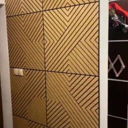 Panel ścienny z drewna o geometrycznym wzorze, tworzącym iluzję głębi. Widoczny fragment szafy wnękowej i włącznik światła, co sugeruje funkcjonalność i estetykę.