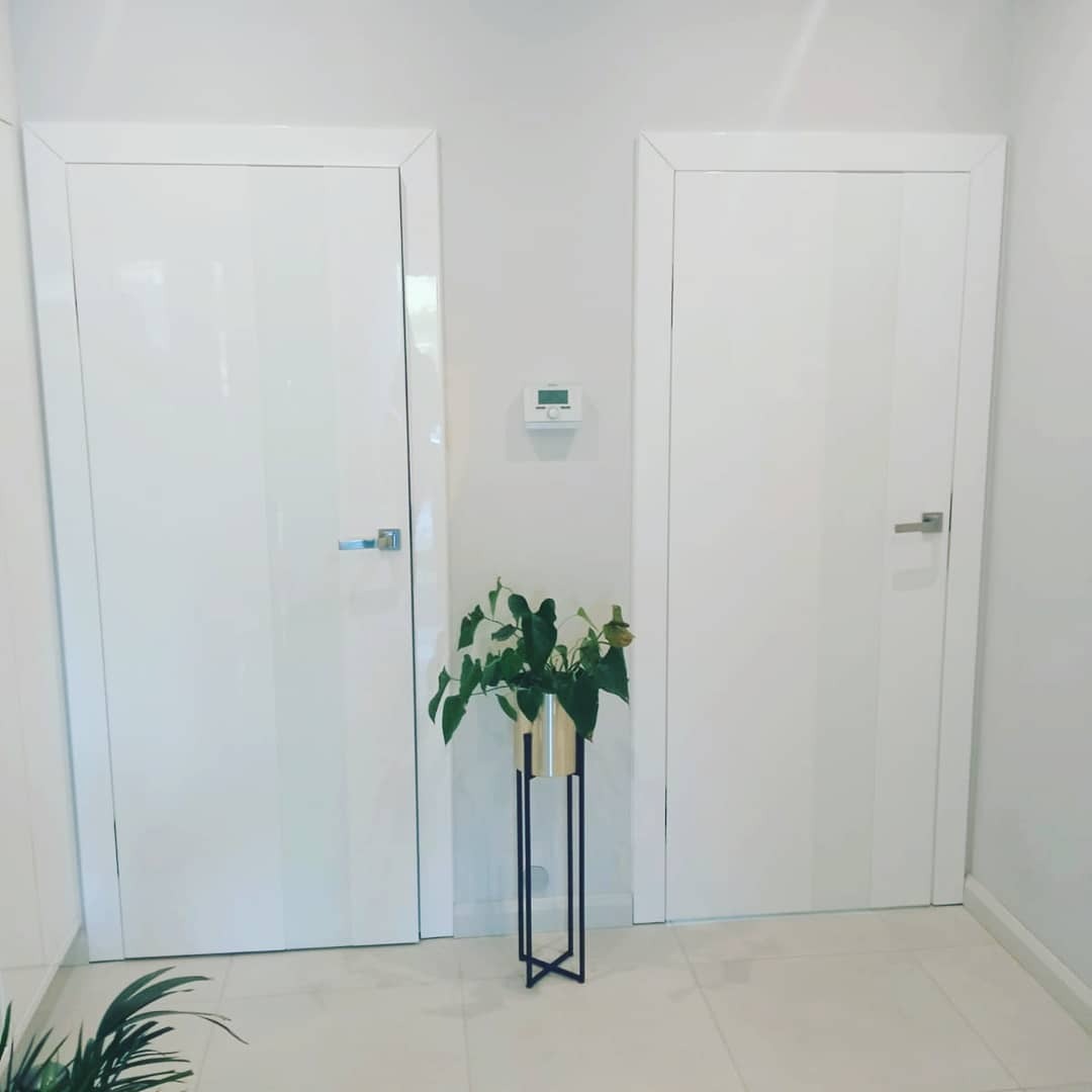 Dwa białe, lakierowane drzwi wewnętrzne z srebrnymi klamkami w korytarzu w Sasino. Pomiędzy nimi roślina w złotej doniczce na czarnym stojaku.