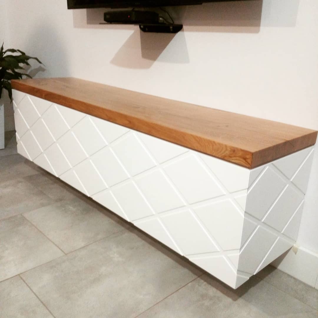 Biała, wisząca szafka RTV z drewnianym blatem i dekoracyjnym frezowaniem w romby, zamontowana na ścianie w nowoczesnym salonie, widok z perspektywy podłogi.