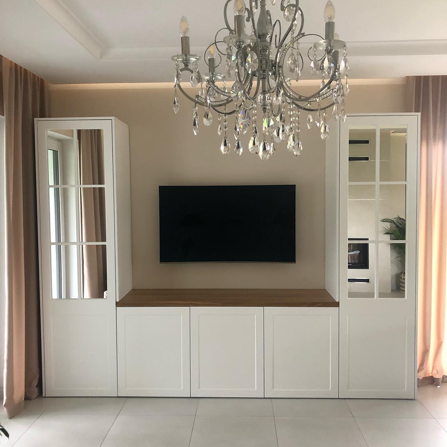 Biała zabudowa meblowa pod telewizor z przeszklonymi witrynami po bokach, drewniany blat, elegancki żyrandol kryształowy, jasne zasłony i płytki podłogowe.