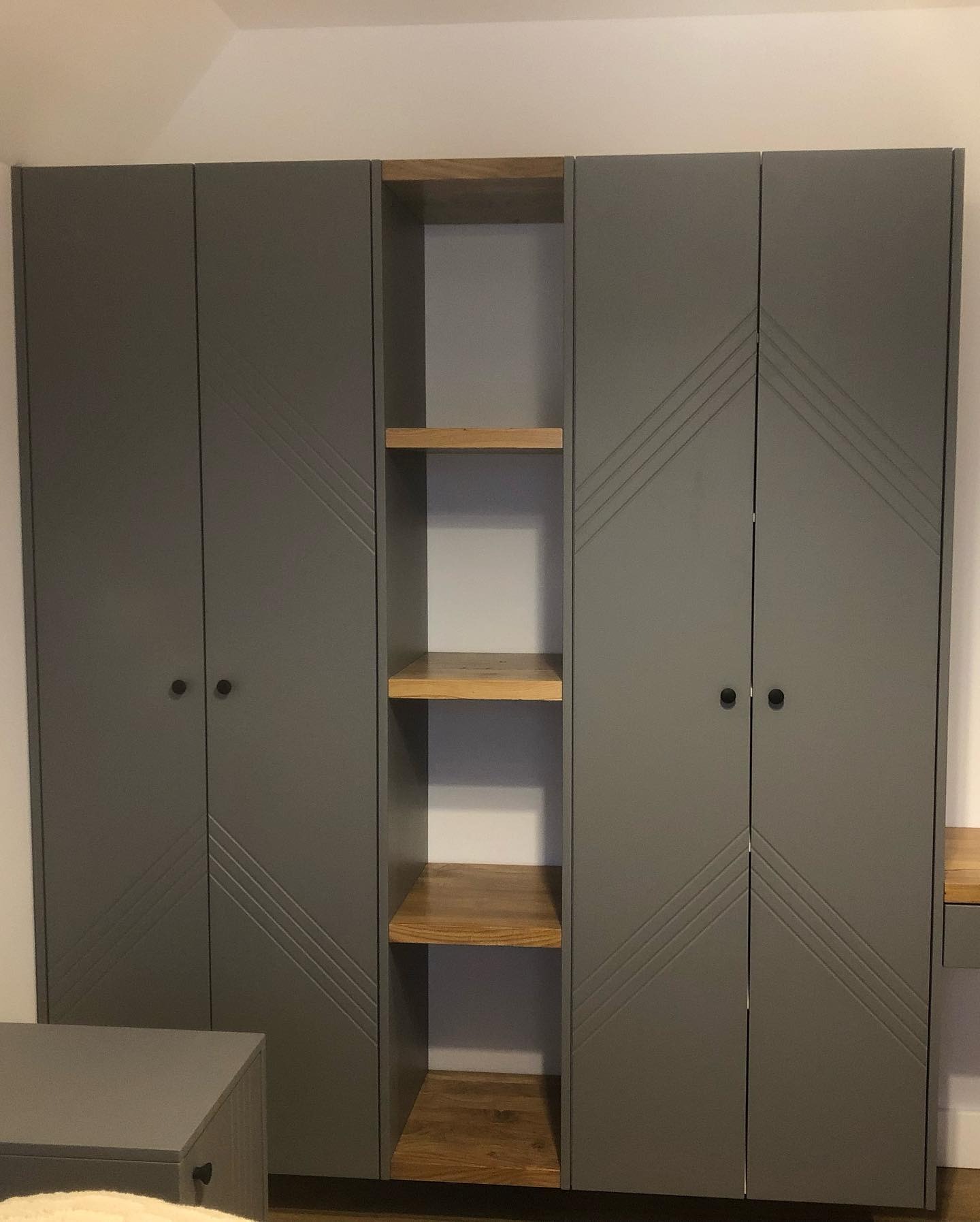 Szara szafa wnękowa z drewnianymi półkami, minimalistyczny design z frezowanymi liniami na frontach, czarne okrągłe uchwyty, oświetlenie górne.