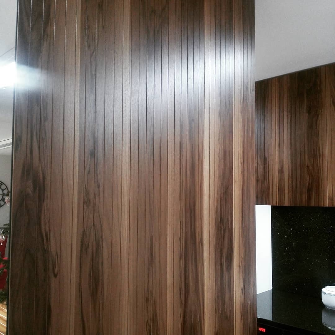 Panel ścienny z drewna o pionowym układzie słojów w odcieniach brązu, tworzący elegancką zabudowę w kuchni z elementami czarnego blatu i białych frontów.