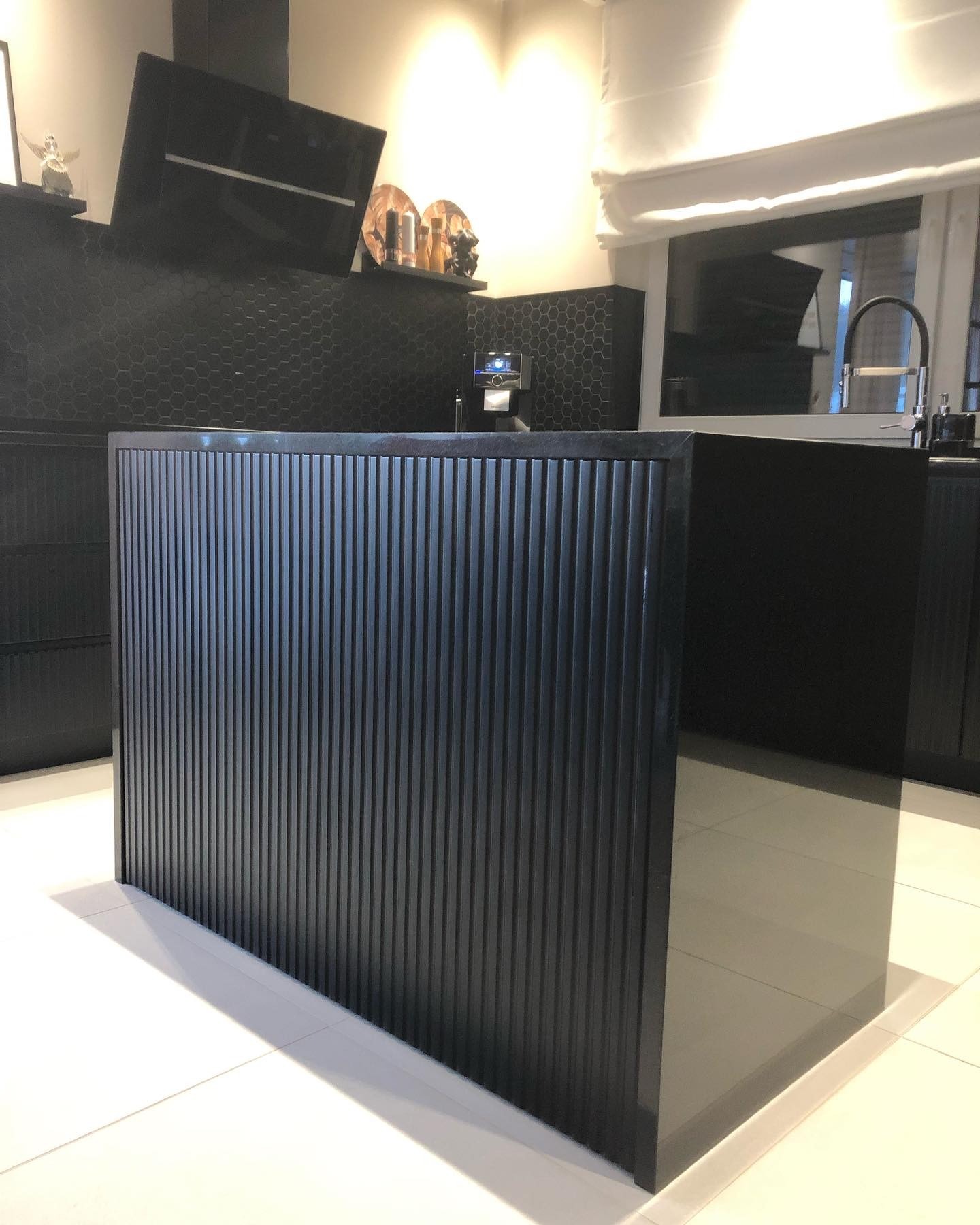 Ciemna wyspa kuchenna z ryflowanym frontem, na białej podłodze, w tle czarne heksagonalne płytki i okno. Minimalistyczny design, nowoczesne wykończenie.