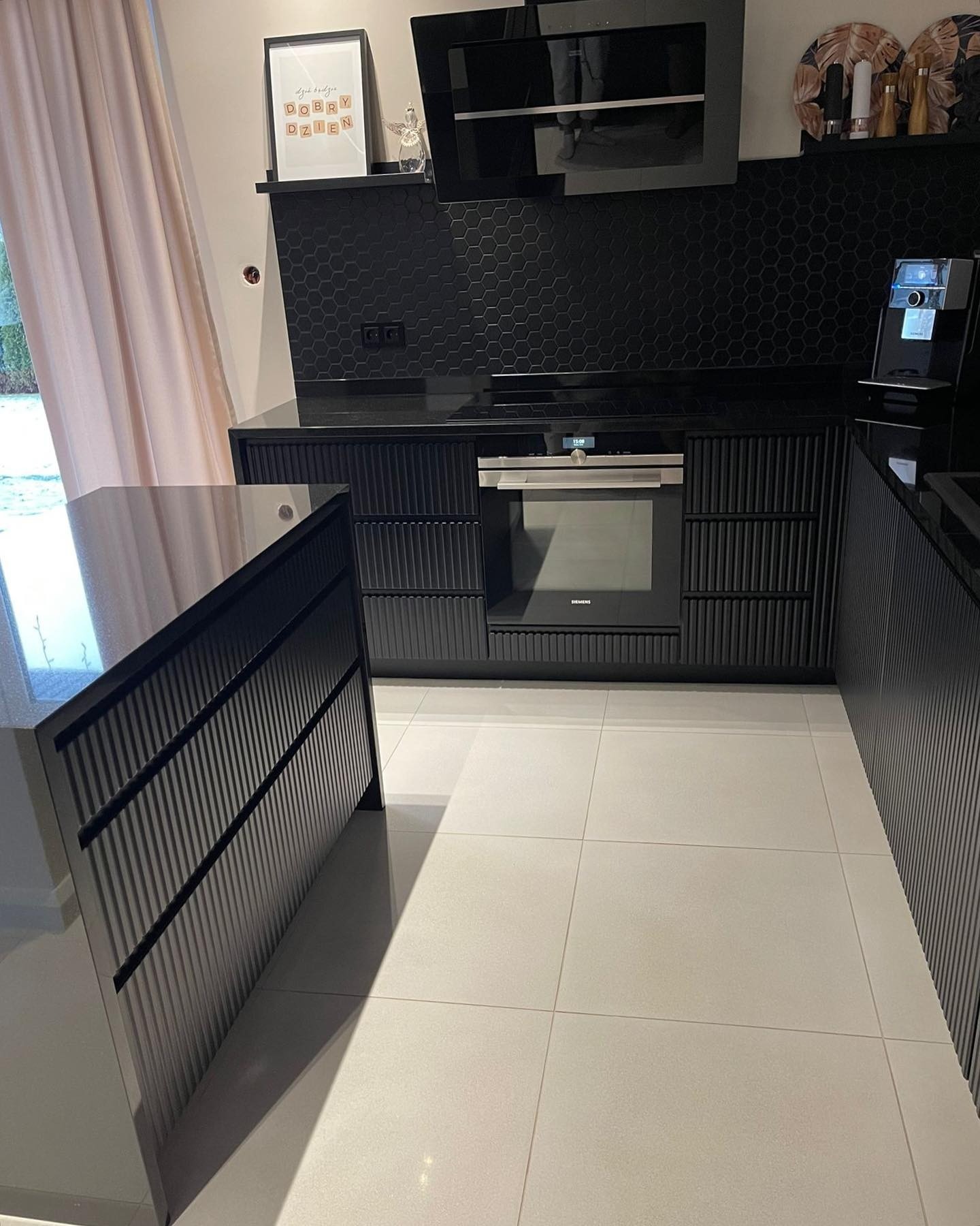 Nowoczesna, czarna kuchnia z frezowanymi frontami, blatem z połyskiem i heksagonalnymi płytkami na ścianie. Widoczny piekarnik Siemens i ekspres do kawy.
