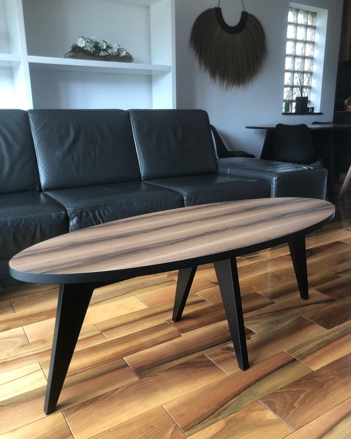 Stylowy, owalny stolik kawowy z drewnianym blatem i czarnymi nogami na tle skórzanej sofy i drewnianej podłogi. Dekoracja ścienna nadaje wnętrzu charakteru.