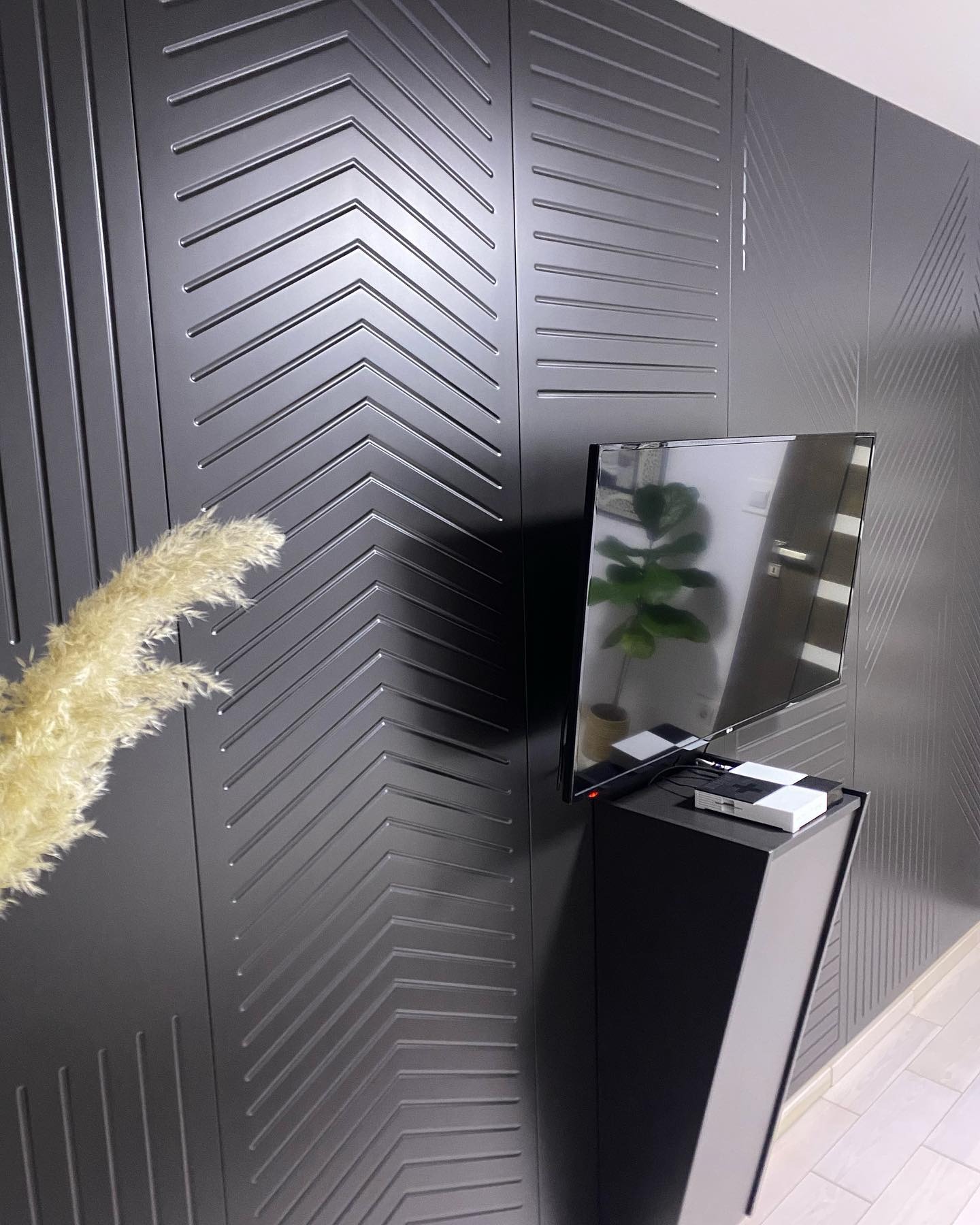 Ciemna ściana z geometrycznym wzorem w jodełkę, na której wisi telewizor na czarnej, smukłej szafce. Obok widoczna trawa pampasowa.