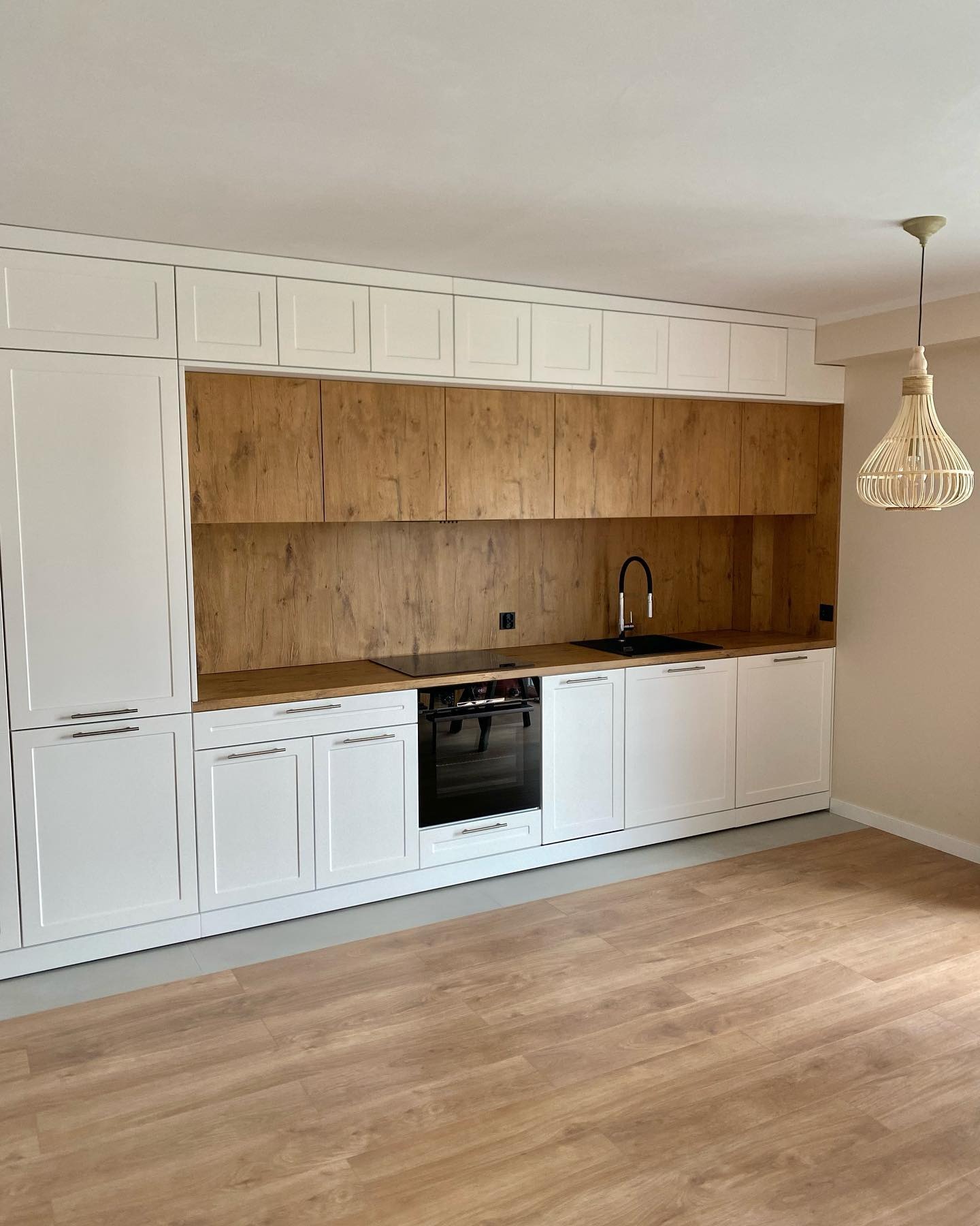 Nowoczesna kuchnia z białymi szafkami dolnymi i górnymi w kolorze drewna, czarny zlew i płyta grzewcza, wisząca lampa z abażurem z wikliny.