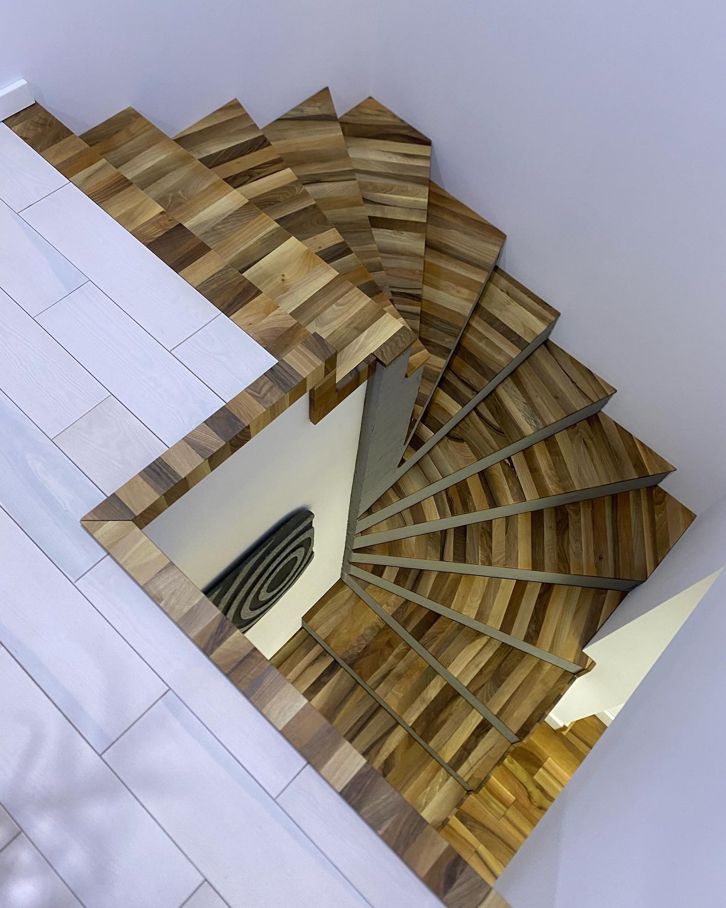 Spiralne schody drewniane z mozaikową fakturą stopni, widok z góry, kontrastujące z białymi panelami podłogowymi i ścianami. Unikalny wzór i nowoczesny design.