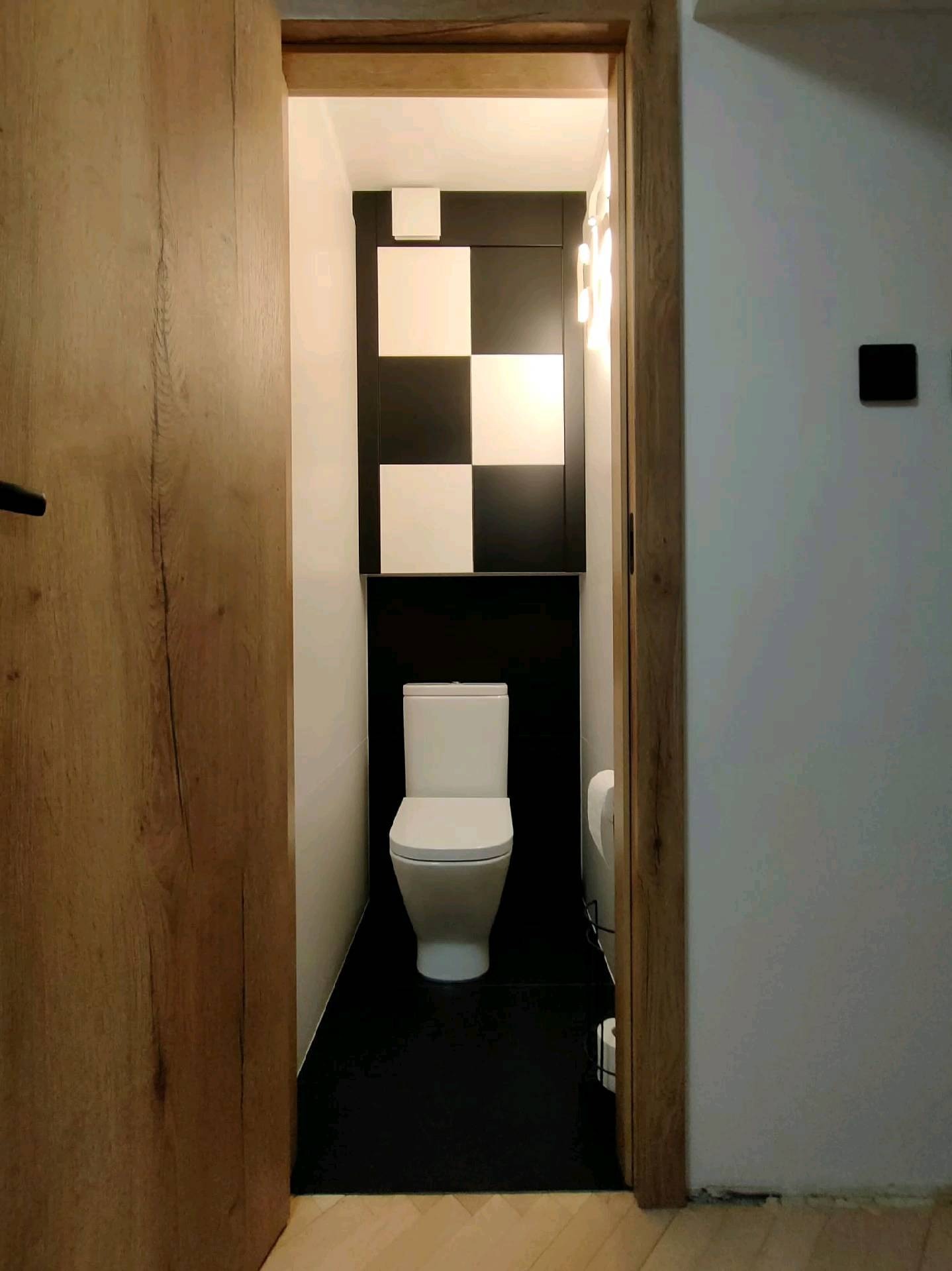 Nowoczesna toaleta z czarno-białą szachownicą nad zabudową WC, widoczna przez otwarte, drewniane drzwi. Minimalistyczny design z akcentem na kontrast kolorów.
