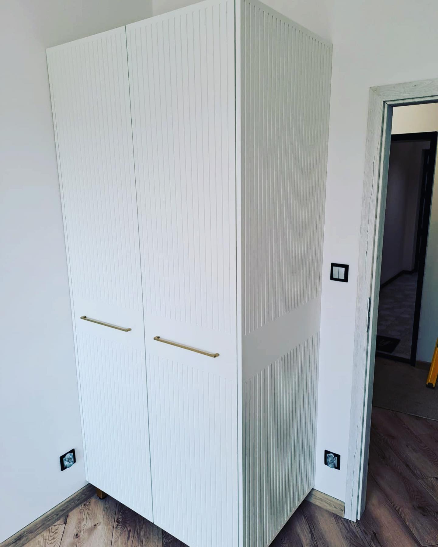 Biała szafa dwudrzwiowa z ryflowanymi frontami i złotymi uchwytami, stojąca w rogu pokoju z drewnianą podłogą. Widoczne gniazdka elektryczne i fragment otwartych drzwi.