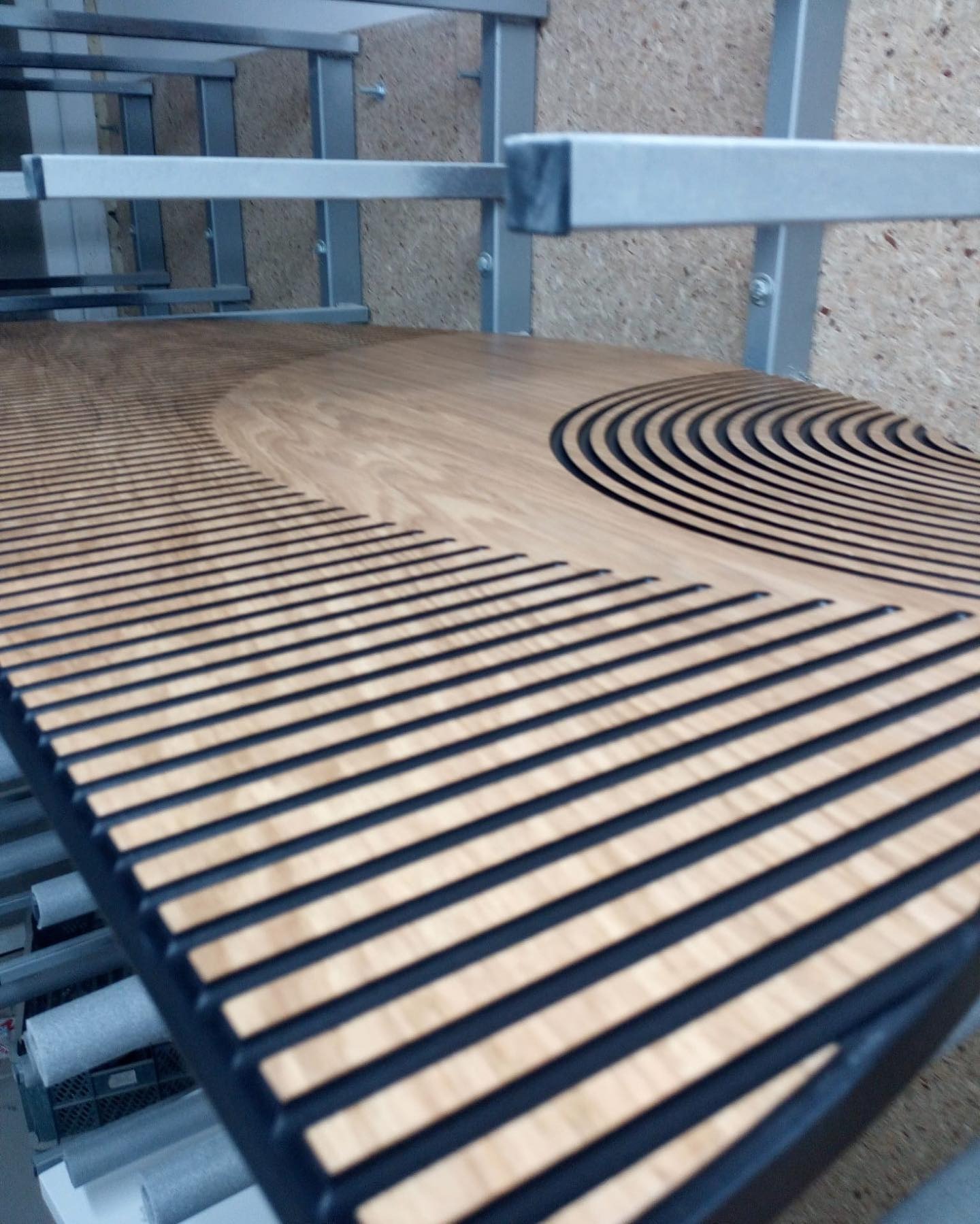 Panel akustyczny z drewna z wyfrezowanymi, równoległymi rowkami i motywem półkola, leżący na metalowym regale w warsztacie. Widok z góry.