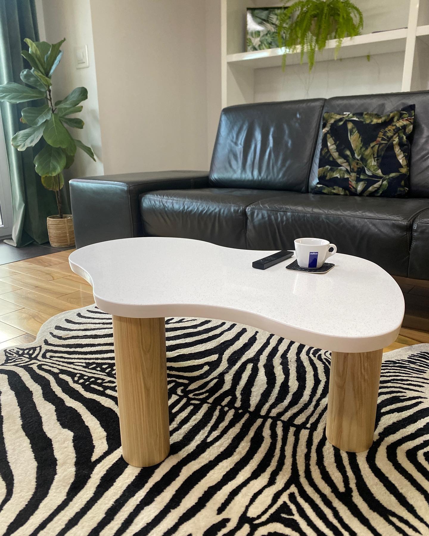 Biały stolik kawowy o nieregularnym kształcie na drewnianych nogach, z filiżanką i pilotem, na tle sofy i dywanu w zebrę. Nowoczesny design.