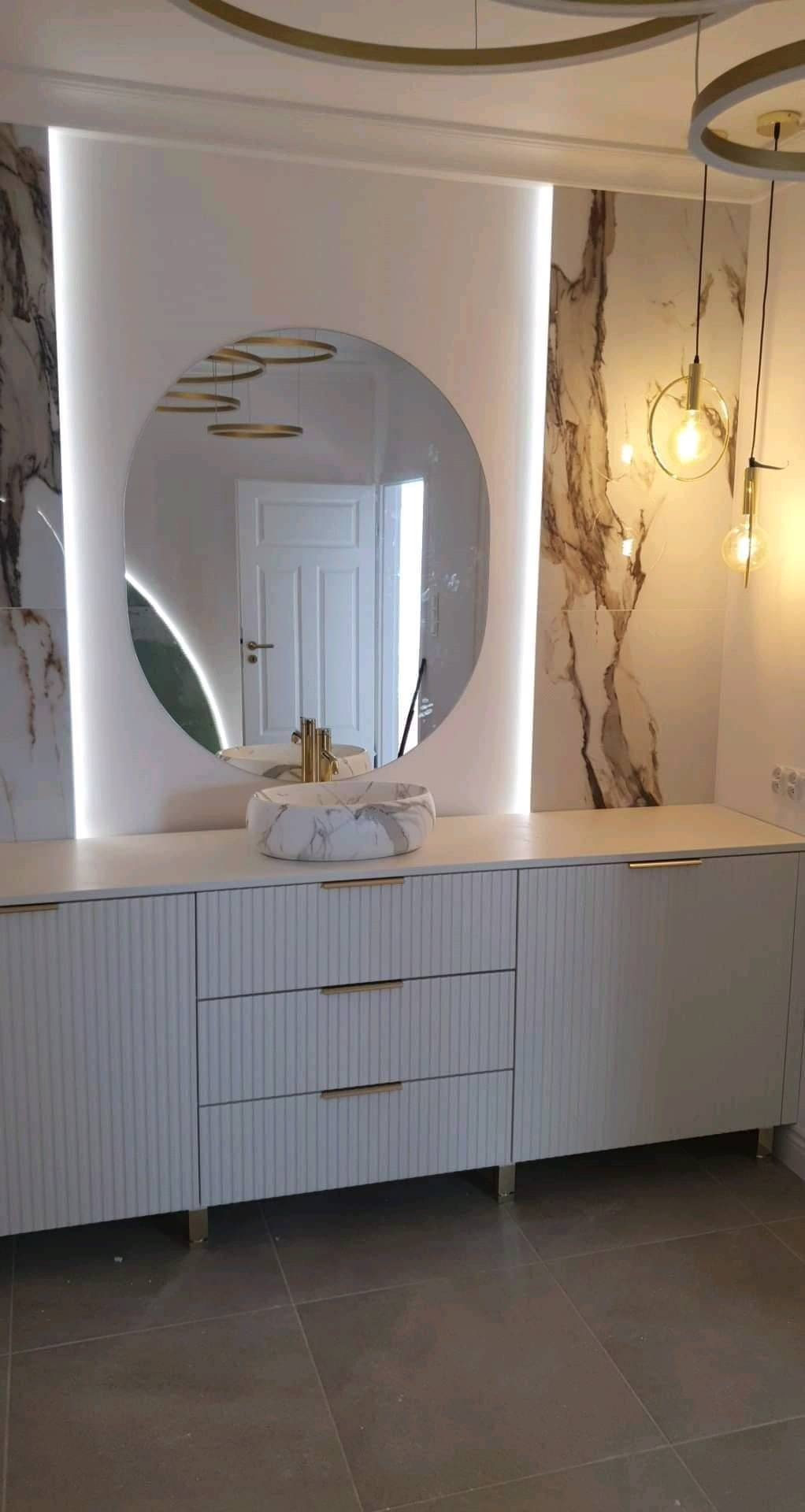 Elegancka łazienka z umywalką w marmurkowy wzór, owalnym lustrem podświetlonym LED i szafką z ryflowanymi frontami oraz złotymi uchwytami. Całość w jasnej tonacji.
