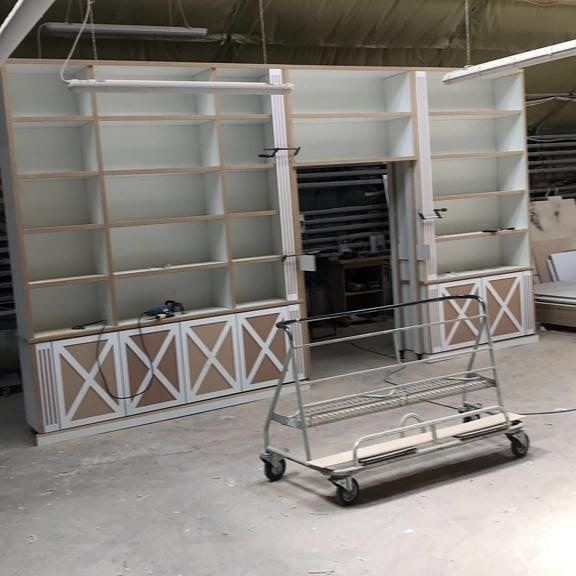 Biała szafa wnękowa z dekoracyjnymi krzyżami na dolnych szafkach, w trakcie montażu. W tle wózek transportowy i elementy warsztatu.