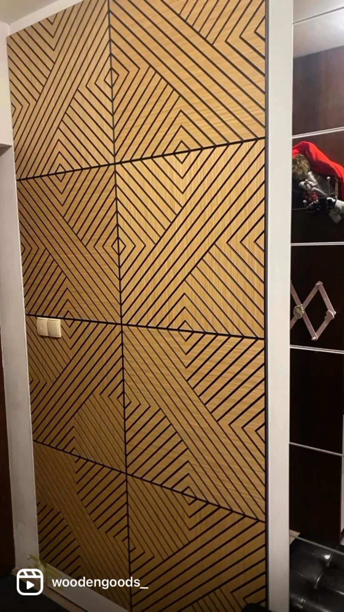 Panel ścienny z drewna o geometrycznym wzorze, tworzącym iluzję głębi. Widoczny fragment szafy wnękowej i włącznik światła, co sugeruje funkcjonalność i estetykę.