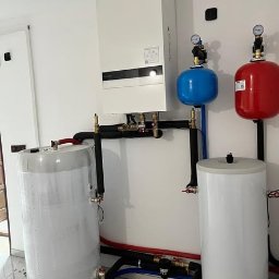 TERMAG SPÓŁKA Z OGRANICZONĄ ODPOWIEDZIALNOŚCIĄ - Nowoczesna instalacja grzewcza z białym kotłem gazowym, dwoma zbiornikami i rurami w kolorach czarnym, niebieskim i czerwonym na tle białej ściany.