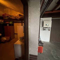TERMAG SPÓŁKA Z OGRANICZONĄ ODPOWIEDZIALNOŚCIĄ - Widok na garaż z zamontowanym inwerterem fotowoltaicznym na ścianie, widoczne przewody i instalacja elektryczna. Częściowo widoczny regał z różnymi przedmiotami.