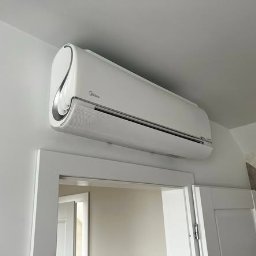 TERMAG SPÓŁKA Z OGRANICZONĄ ODPOWIEDZIALNOŚCIĄ - Biały klimatyzator Midea, zamontowany nad futryną drzwi, widoczny z dołu pod kątem. Minimalistyczny design, jasne wnętrze, subtelne oświetlenie.