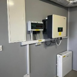 TERMAG SPÓŁKA Z OGRANICZONĄ ODPOWIEDZIALNOŚCIĄ - Szara ściana z zamontowaną instalacją fotowoltaiczną: inwerter, skrzynka rozdzielcza i akumulator. Widoczny fragment rury wentylacyjnej pod sufitem.