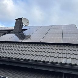 TERMAG SPÓŁKA Z OGRANICZONĄ ODPOWIEDZIALNOŚCIĄ - Panele fotowoltaiczne na dachu domu z dachówką ceramiczną, widoczne komin i antena satelitarna. Pochmurne niebo w tle. Instalacja fotowoltaiczna.